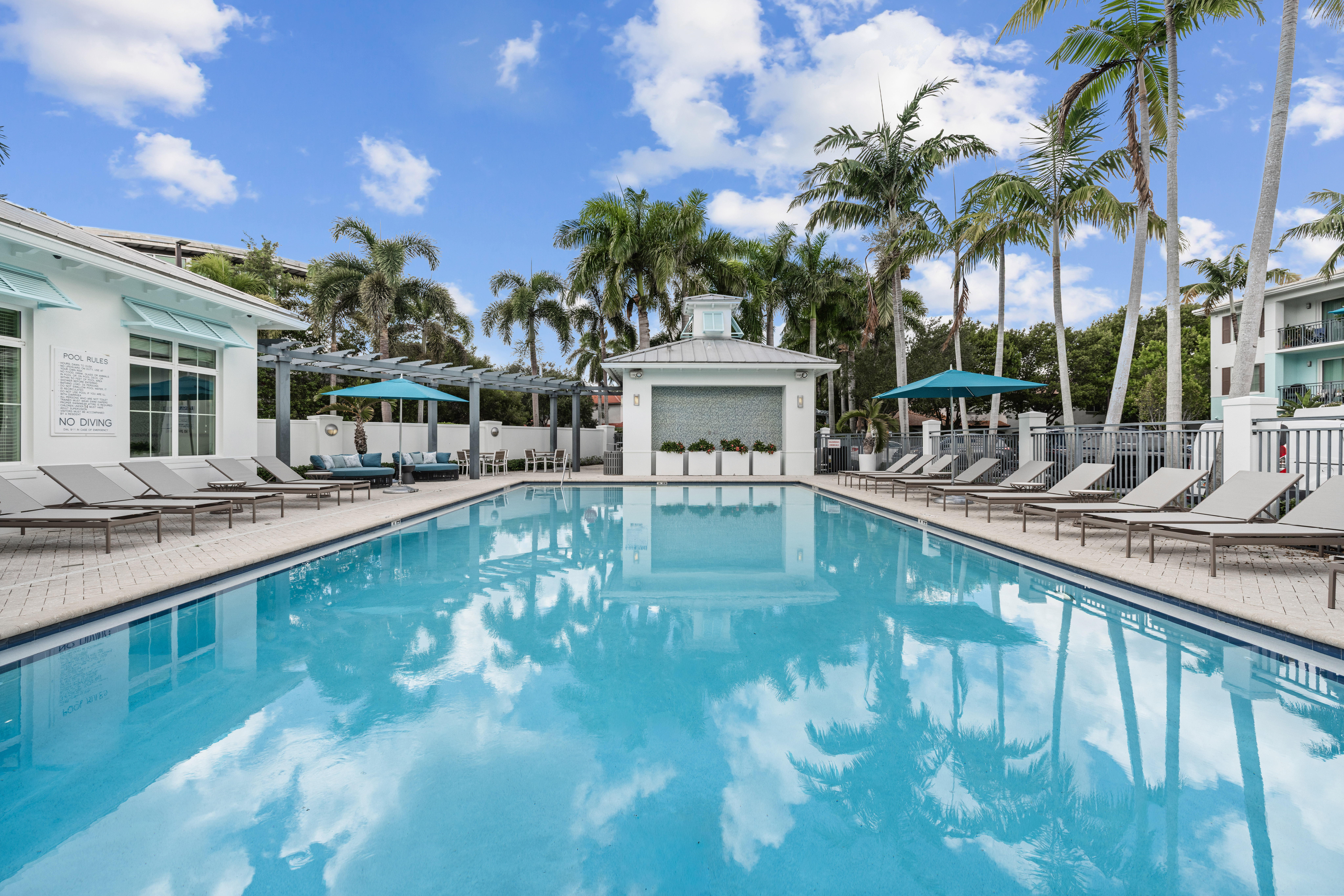 A clear blue swimming pool surrounded by lounge chairs, umbrellas, palm trees, and modern white buildings under a partly cloudy sky.