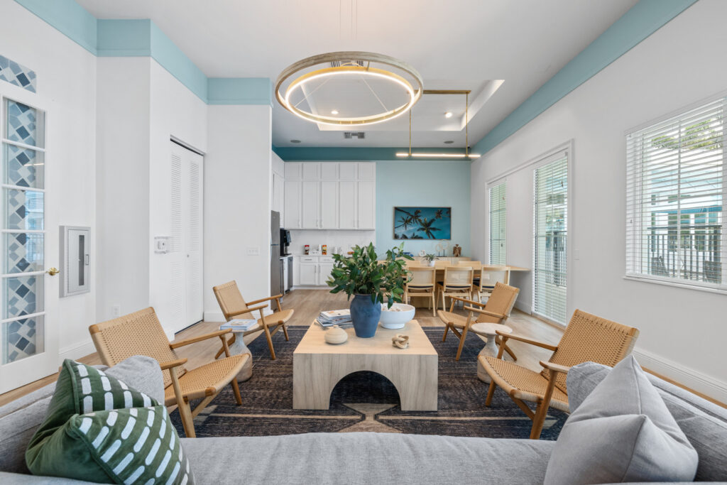 A modern living room with light wood chairs, a gray sofa, a rectangular coffee table with plants, large windows, and white walls with blue trim. A circular ceiling light hangs above the seating area.