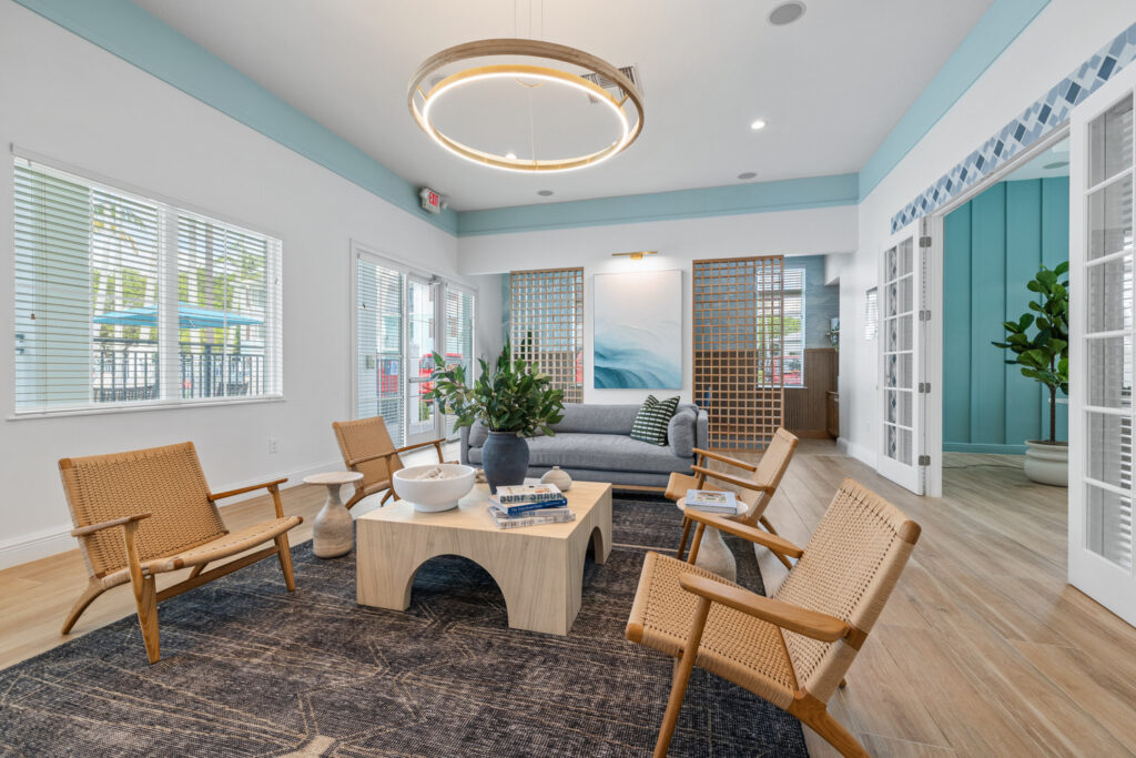A modern, bright lounge features four wooden chairs around a light coffee table, a gray sofa, large circular ceiling light, and coastal decor. Large windows and glass doors let in natural light, with plants and artwork adding warmth.