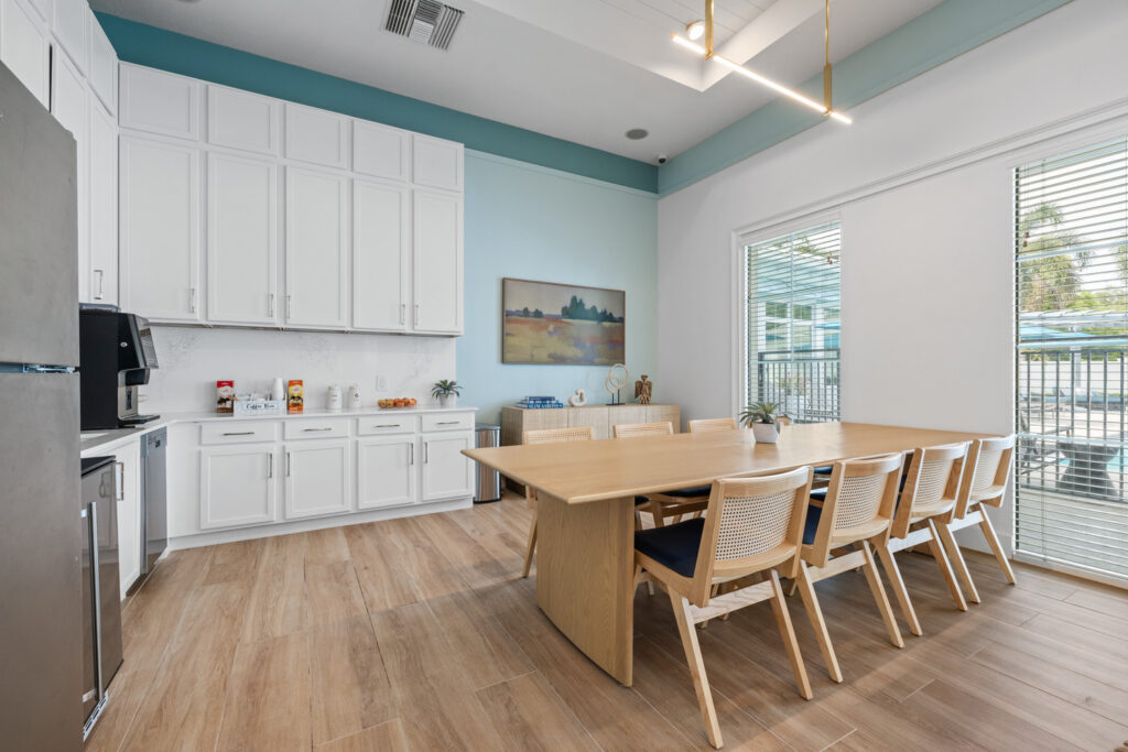 A modern kitchen with white cabinets, a wooden dining table with eight chairs, large windows with blinds, and decorative artwork on the pale blue wall. Light streams in, illuminating the clean, spacious room.