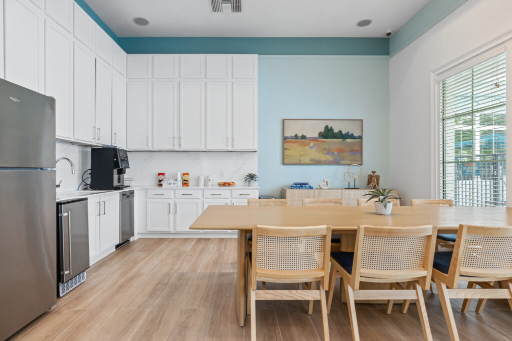 Modern kitchen and dining area with white cabinets, stainless steel appliances, light wood floors, a long wooden table with six chairs, potted plants, a large window, and a colorful landscape painting on a light blue wall.