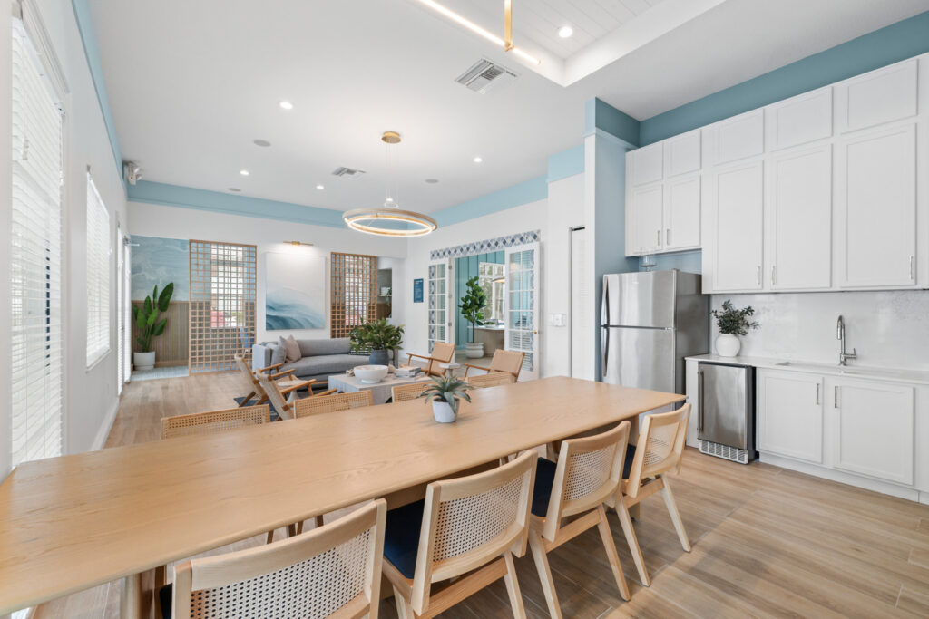 Modern open-concept kitchen and living area with white cabinets, stainless steel appliances, a long wooden dining table, light wood floors, and stylish, minimalist decor with plants and natural light.