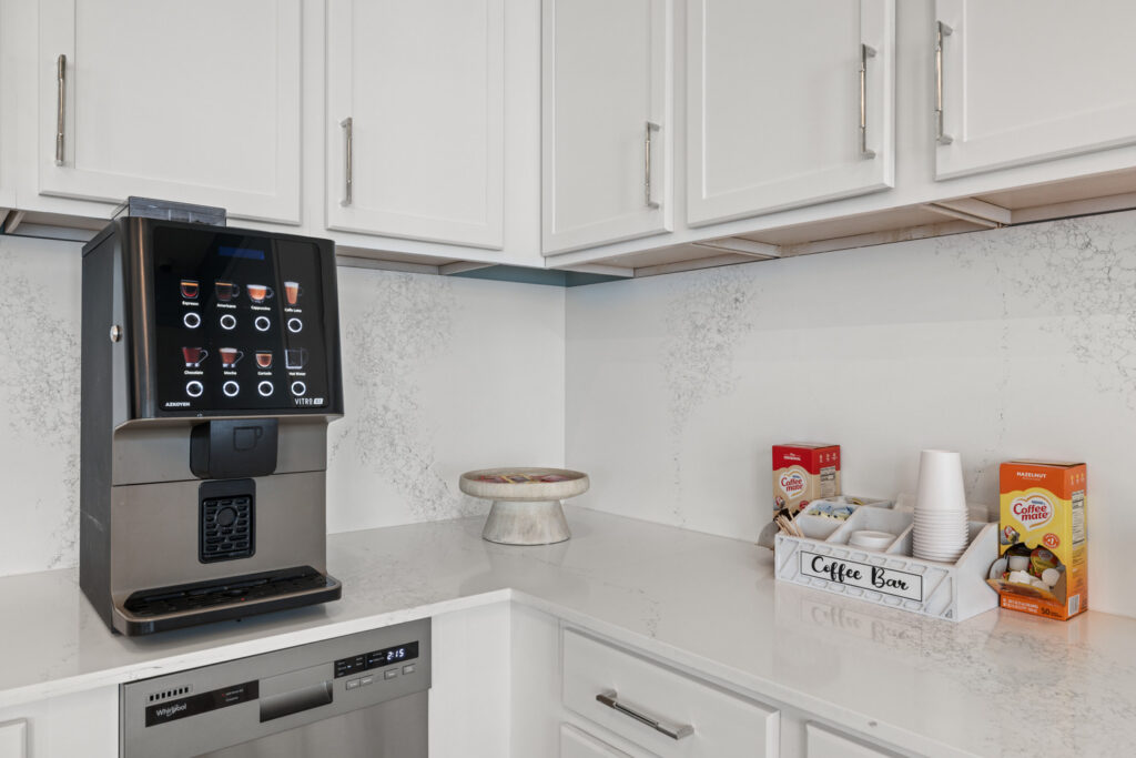 A modern coffee station with a sleek coffee machine, a tray labeled Coffee Bar holding cups and supplies, and boxes of coffee mix on a white countertop with white cabinets above.