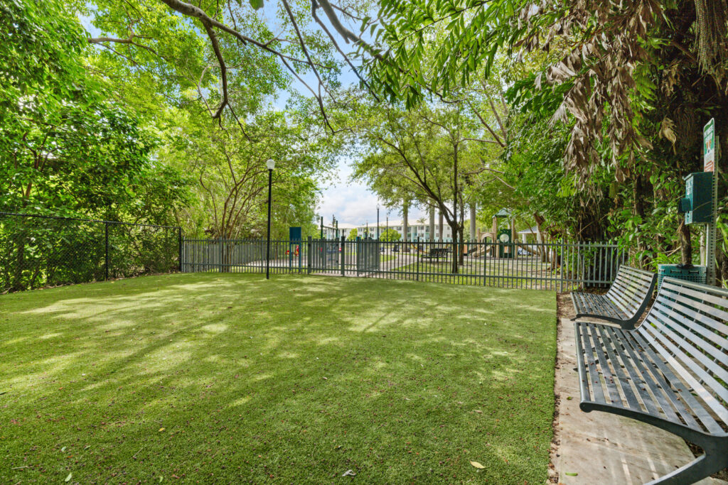 A fenced dog park with green artificial turf, shaded by leafy trees. There are black metal benches along the right side, a sidewalk, and a gated entrance in the background. Buildings and a blue sky are visible beyond the fence.