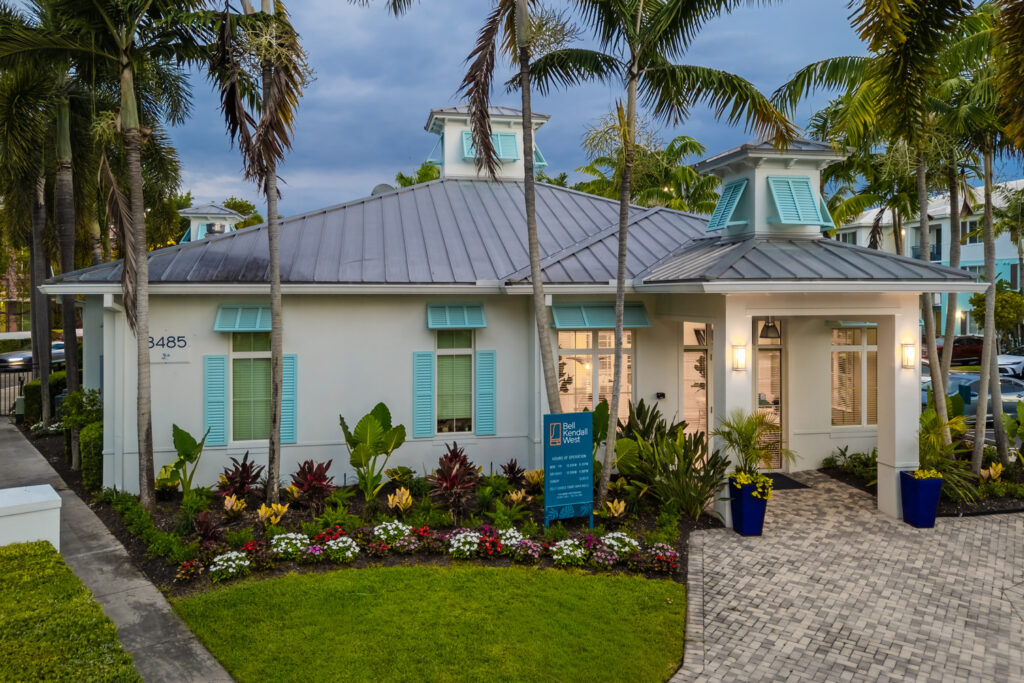 A light blue and white building with a metal roof, surrounded by palm trees, lush landscaping, and colorful flowers; a blue sign stands near the entrance on a paved walkway.