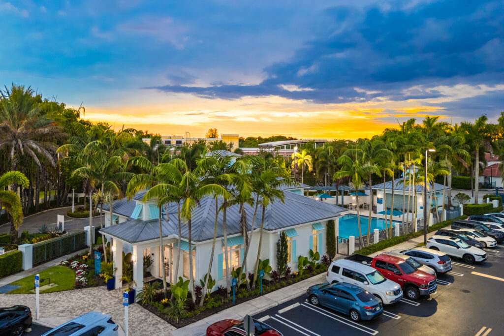 A modern building surrounded by palm trees with a swimming pool, parked cars, and a dramatic sunset sky in the background. The scene appears to be part of a resort or residential community.