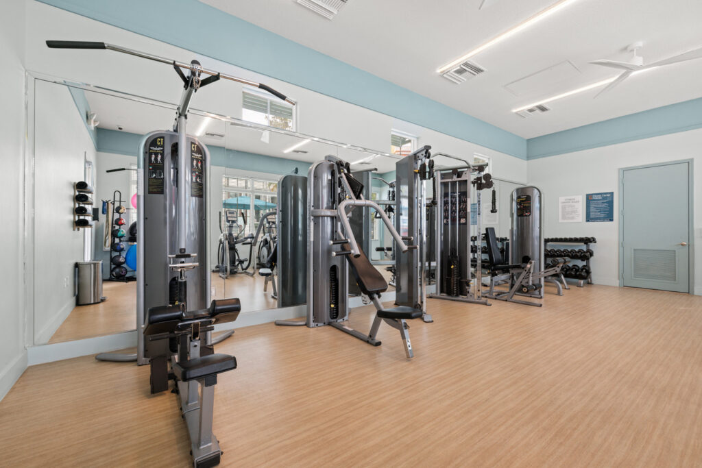 A modern gym with strength training machines, free weights, mirrors on the walls, and bright overhead lighting. The floor has a wood-like finish, and the walls are light blue and white.
