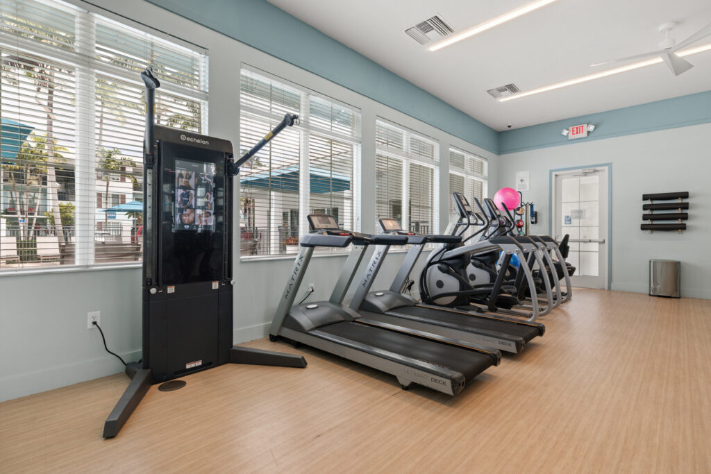 A modern gym with large windows, three treadmills, an elliptical machine, a fitness mirror station, a rack with exercise mats, and a trash can on a light wood floor.