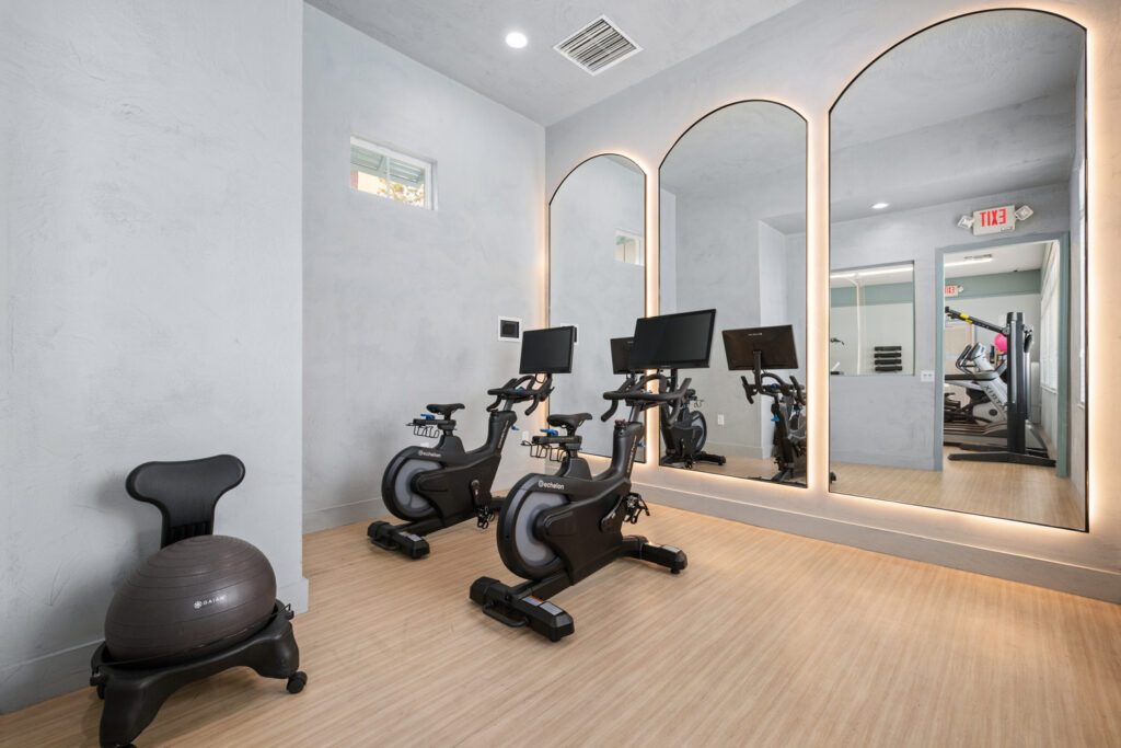 A modern home gym with two stationary bikes facing large mirrored panels, a gray stability ball with a base on the left, light wood flooring, and pale gray walls.