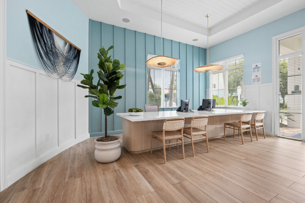 Modern office with light blue walls, wood flooring, and a large desk with four chairs. Two computers sit on the desk, beneath pendant lights. A potted plant and abstract wall art add decorative touches. Large windows let in sunlight.