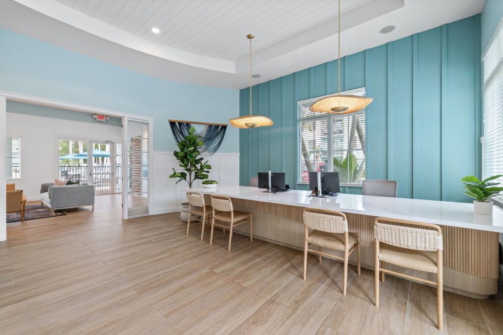Modern office space with a long white desk, four chairs, two computers, pendant lights, and large windows. Light wood floors, teal accent wall, and plants create a bright, welcoming atmosphere.