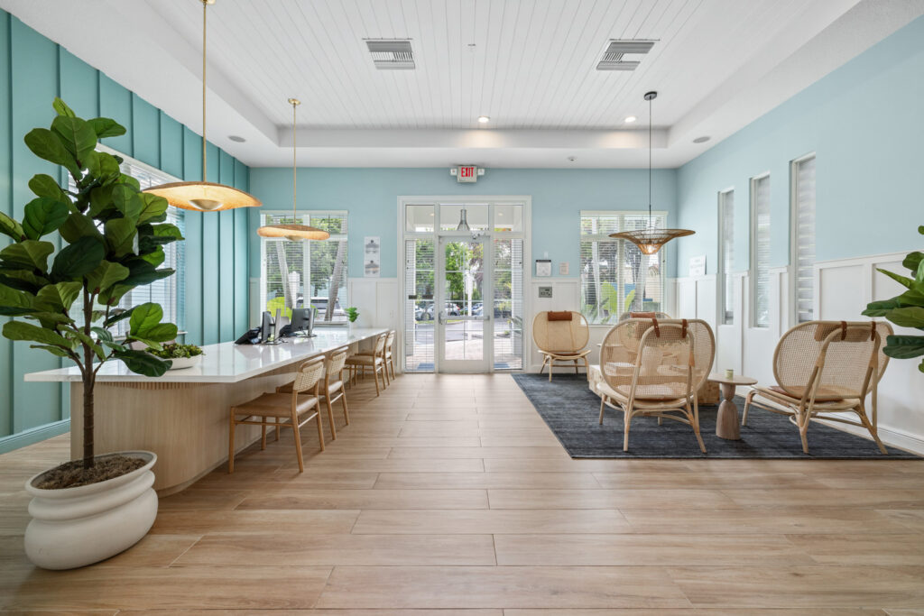 Bright, modern lobby with light wood floors, blue walls, large potted plants, desk with chairs on the left, and cozy seating area with wicker chairs on a dark rug near glass double doors.