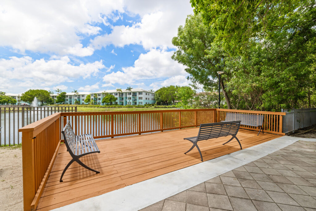 A wooden deck with two metal benches overlooks a pond, with modern apartment buildings and palm trees visible across the water; trees and a partly cloudy sky complete the peaceful outdoor scene.