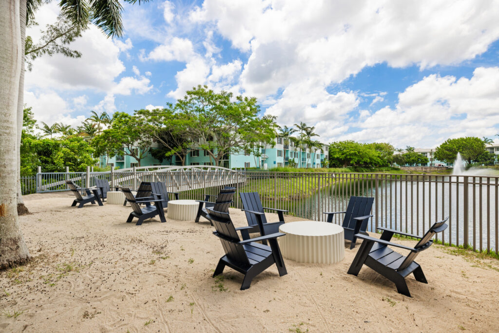 A sandy patio with black Adirondack chairs and round tables overlooks a pond with a fountain, surrounded by trees, a footbridge, and pastel-colored buildings under a partly cloudy sky.
