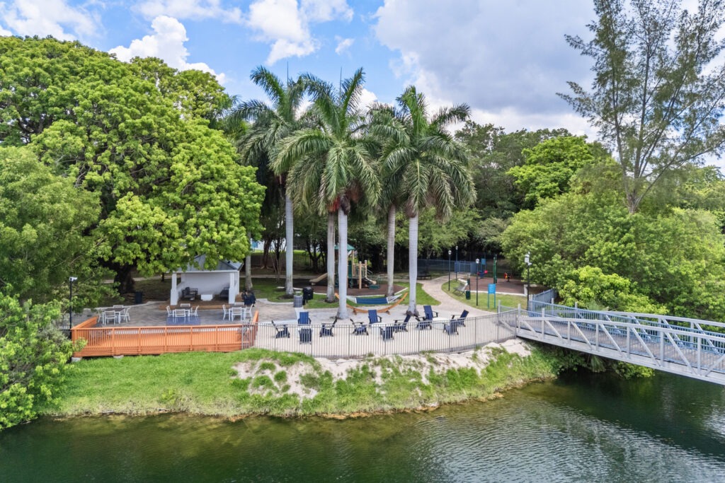 A riverside park with palm trees, outdoor seating, a pavilion, and a metal footbridge over the water, surrounded by lush green foliage under a partly cloudy sky.