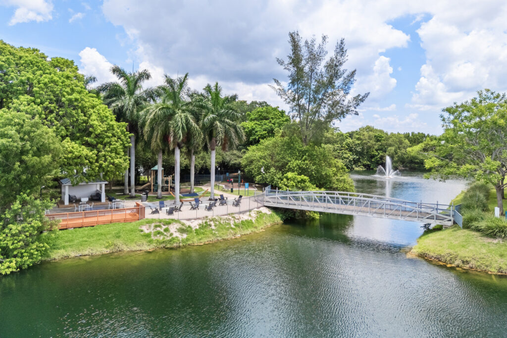 A small bridge crosses over a tranquil lake surrounded by lush green trees, palm trees, and a patio area with seating. A fountain sprays water in the background under a partly cloudy sky.