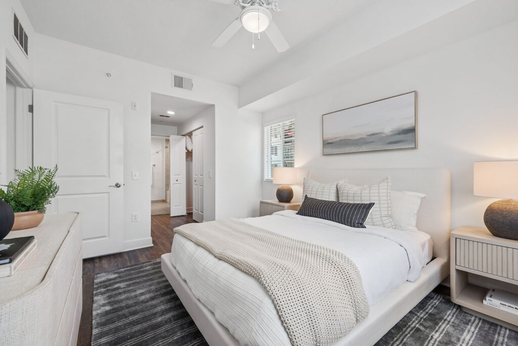 Bright, modern bedroom with a large bed, neutral bedding, two bedside tables with lamps, a dresser with a plant, a window, wall art above the bed, and a gray rug on a wooden floor.
