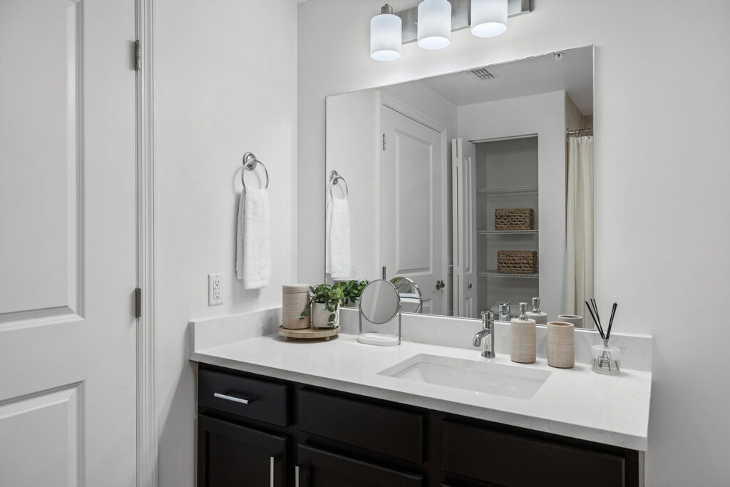 Modern bathroom vanity with a white countertop, dark cabinets, a large mirror, and three lights above. The counter holds towels, a plant, soap, a small mirror, and decorative containers. White walls and door are visible.