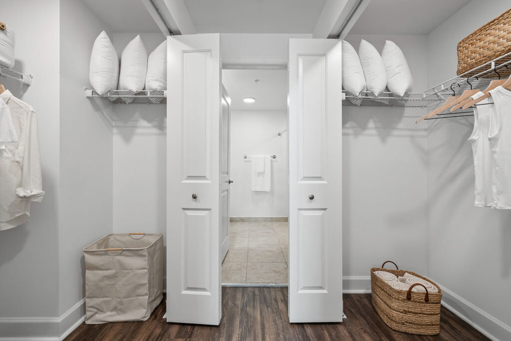 A neatly organized walk-in closet with double white doors, hanging clothes, shelves holding white pillows, a laundry hamper, woven baskets, and a view into a tiled bathroom beyond.