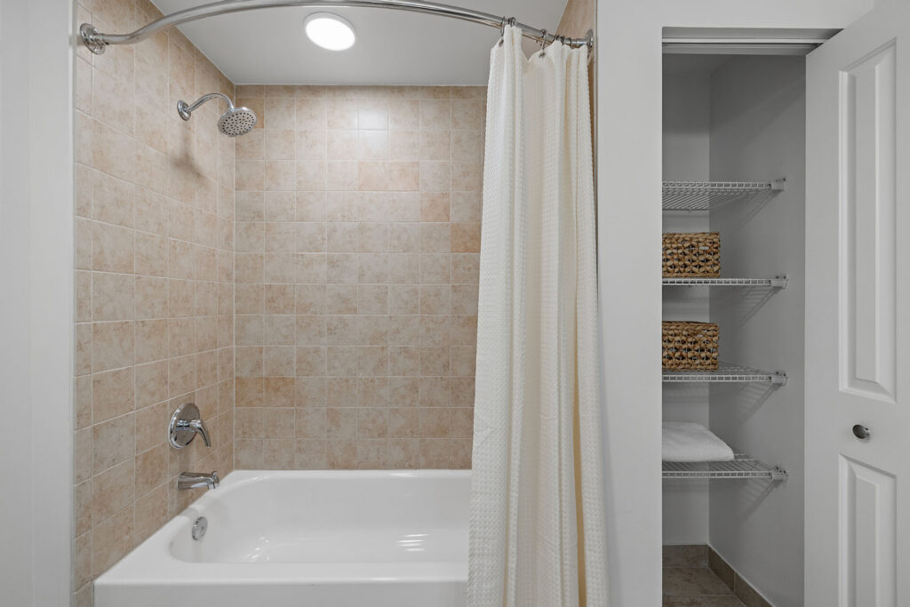 A bathroom with a tiled shower and bathtub, a beige shower curtain, and an open closet with wire shelves holding woven baskets and folded towels.