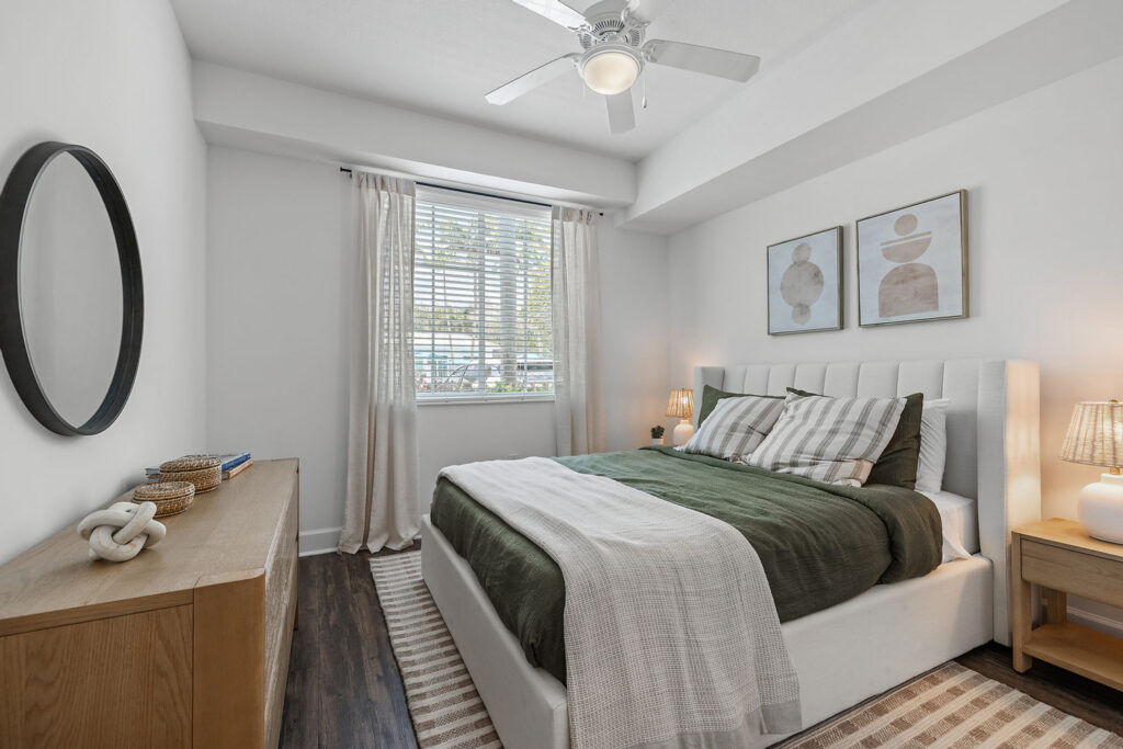 A modern bedroom with a white bed, green and white bedding, two lamps on wooden nightstands, a wooden dresser with decor, a round mirror, and a window with white curtains letting in natural light.