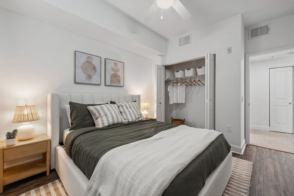 A modern bedroom with a neatly made bed, striped pillows, beige throw blanket, two nightstands with lamps, abstract wall art, and an open closet with hangers and white shirts. The room has white walls and wood flooring.