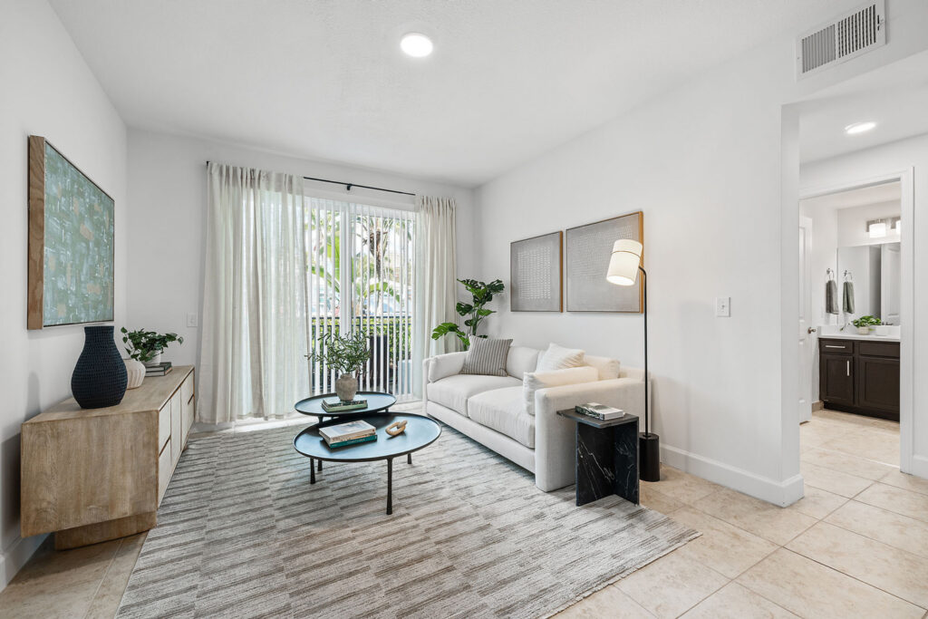A bright, modern living room with a white sofa, round coffee tables, a light rug, and wall art. Large glass doors with sheer curtains let in natural light and reveal greenery outside. A bathroom is visible through an open doorway.