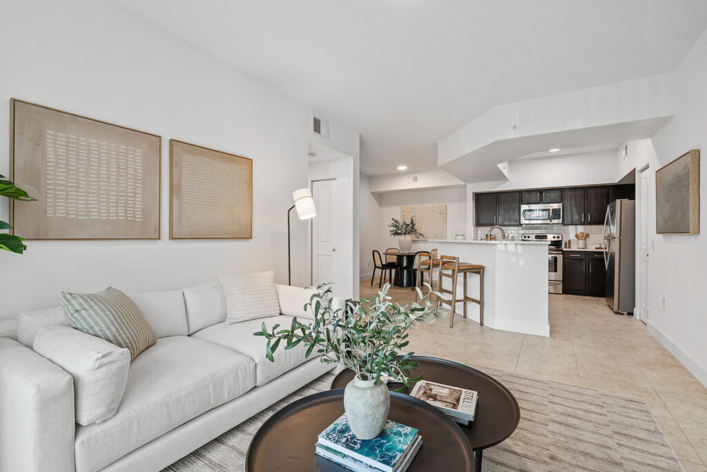 Modern open-concept living room and kitchen featuring a light sofa, coffee tables with books and a plant, neutral wall art, dining table with chairs, and dark wood kitchen cabinets with stainless steel appliances.